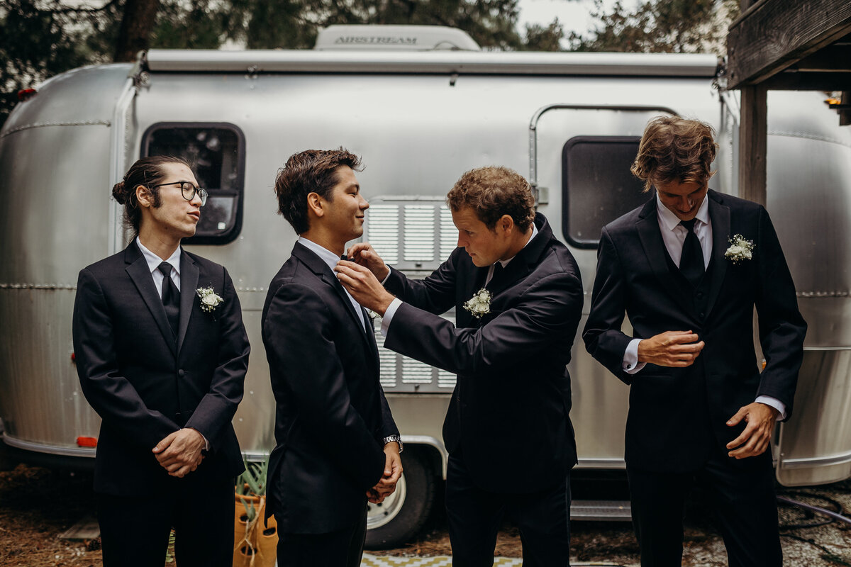 A groomsman adjusts the groom's tie in front of an airstream trailer.