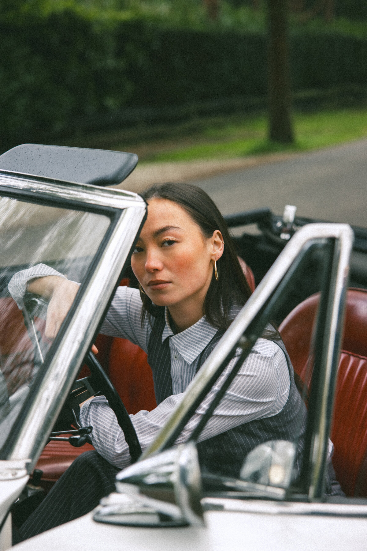 A side view of a woman in a classic car, exuding a cool, collected vibe. Her attire and the car’s design complement each other, evoking a timeless sense of style. The shot is framed beautifully, with the car’s details adding depth to the composition, photographed by Bo Eppink.