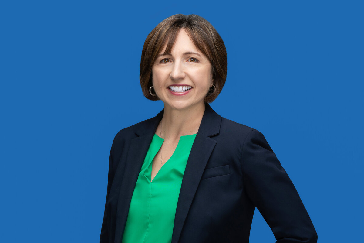Vibrant headshot of a brunette woman in a sharp navy blazer and striking green blouse, set against a bold blue backdrop, showcasing Cincinnati's dynamic professionals.