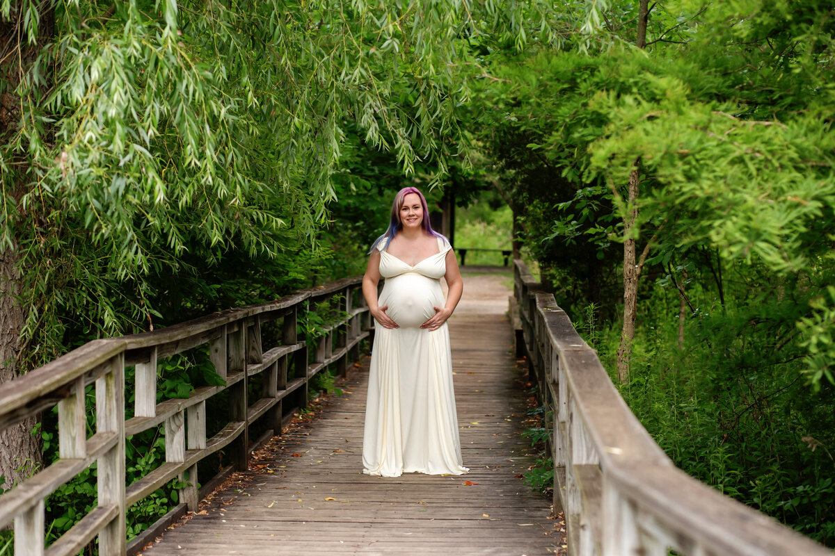 A pregnant woman standing on a wooden bridge smiling at the camera. She's wearing a white dress and has blue and purple in her blonde hair.