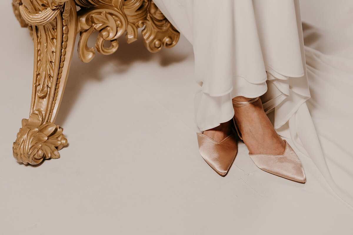 A close-up of the bride’s satin pointed shoes resting on the edge of an ornate gold chair, with the flowing dress cascading around them.