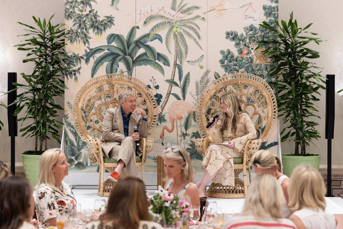 A speaker and moderator seated in woven chairs on stage engaging in dialogue before a seated audience.