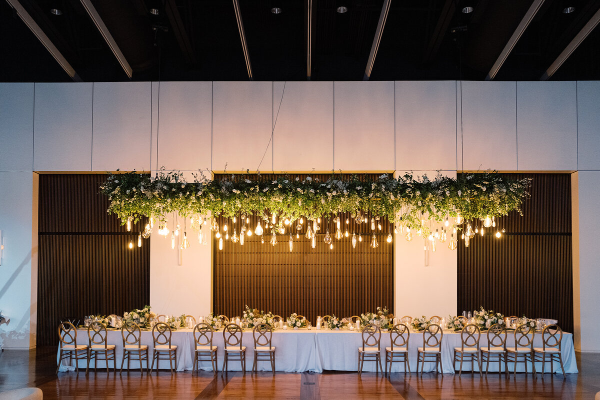 Epic floral installation for garden-inspired summer wedding. Large and lush floral install over head table for timeless wedding design. Vine-like greenery and florals accent this hanging Edison bulb installation. Downtown classic white and green wedding. Design by Rosemary & Finch Floral Design in Nashville, TN.