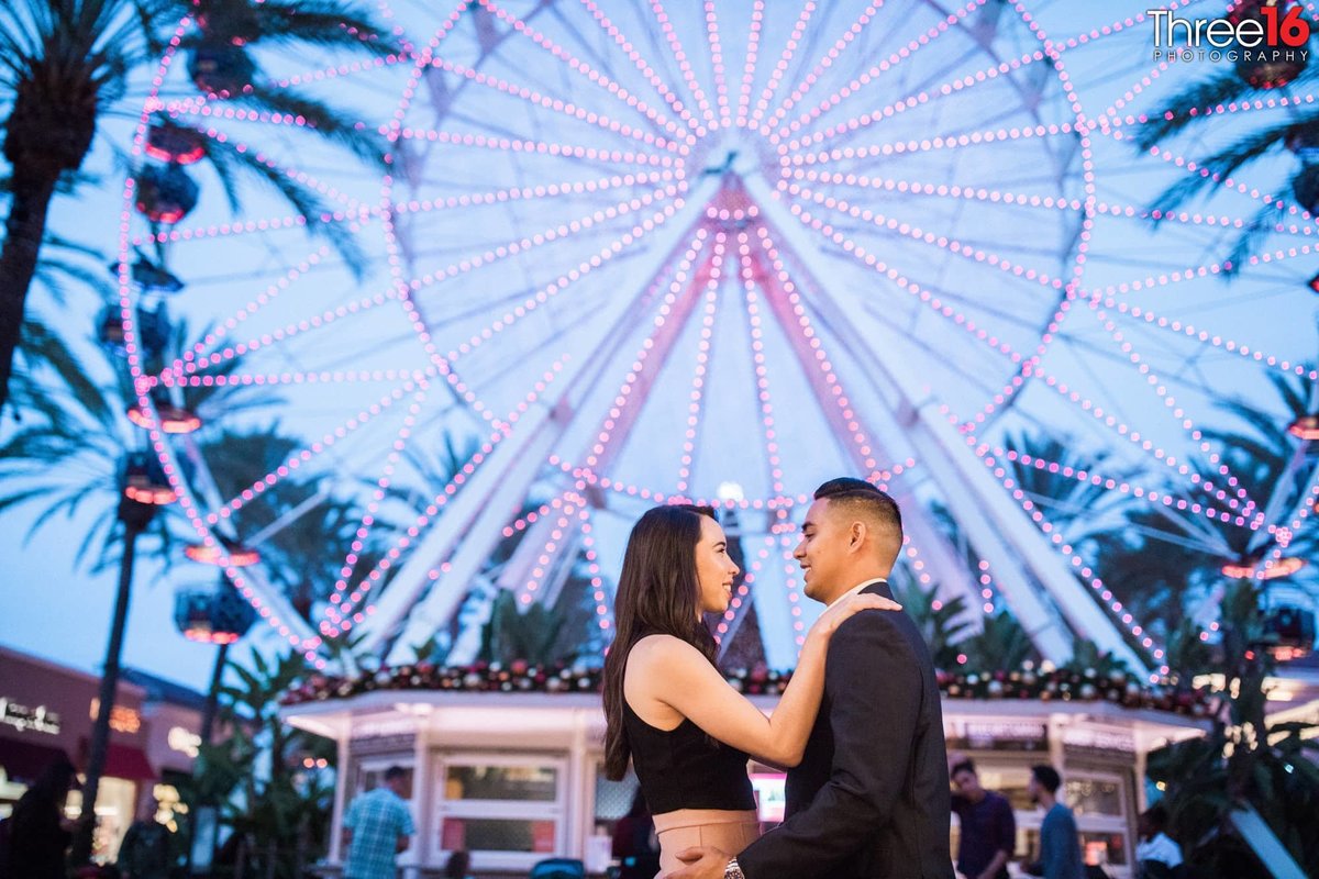 Future Bride and Groom hold each other and talk in front of the Ferris Wheel at the Irvine Spectrum in the evening