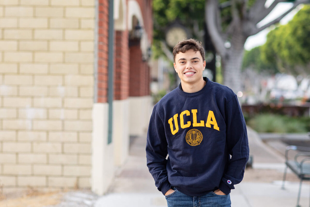 A person wearing a UCLA sweatshirt smiles while standing on a sidewalk, with trees and buildings in the background.