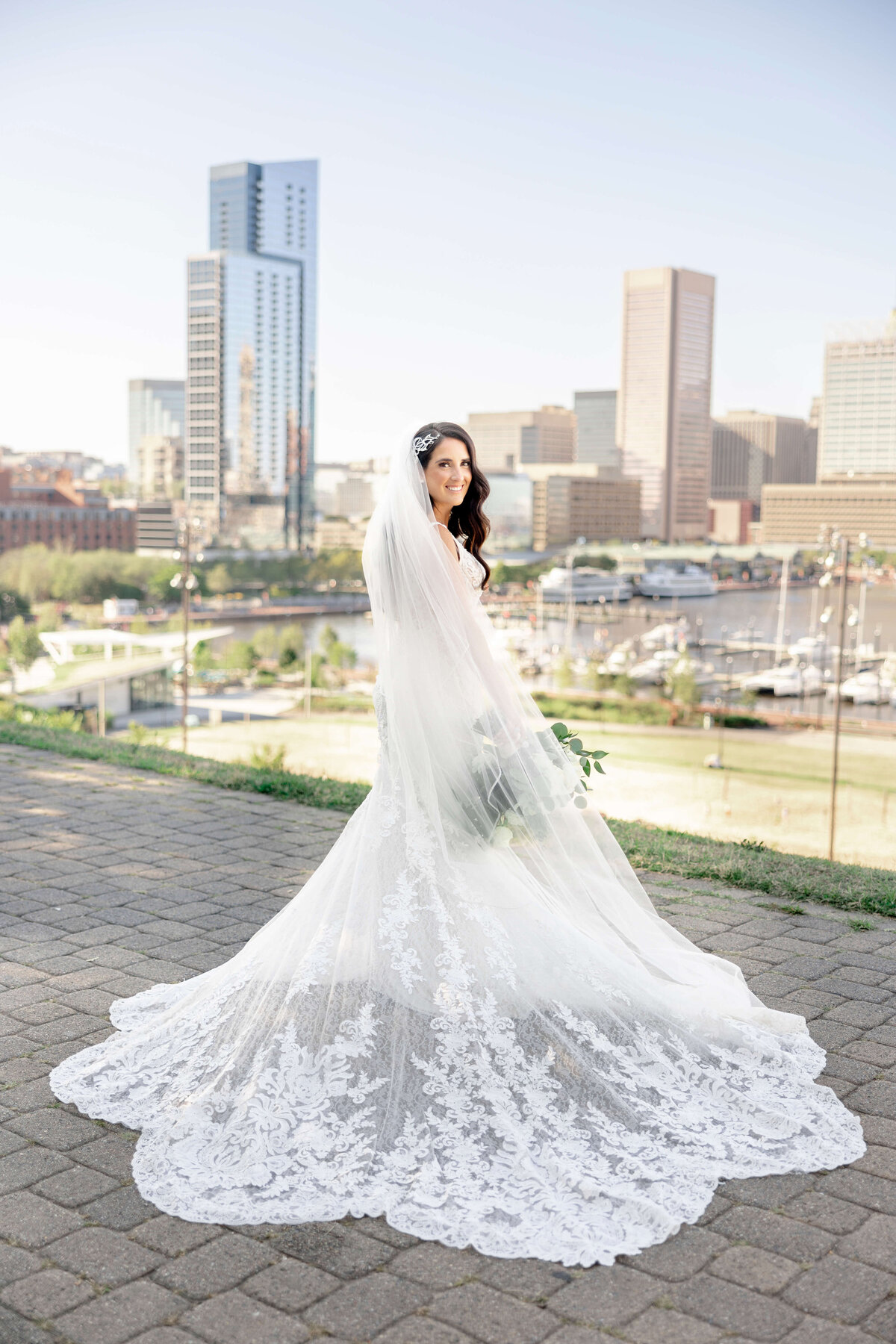A bride in a white lace wedding gown stands on a paved path overlooking a cityscape. Her long veil and train spread gracefully behind her. The hazy skyline and high-rise buildings are visible in the background on a clear day.