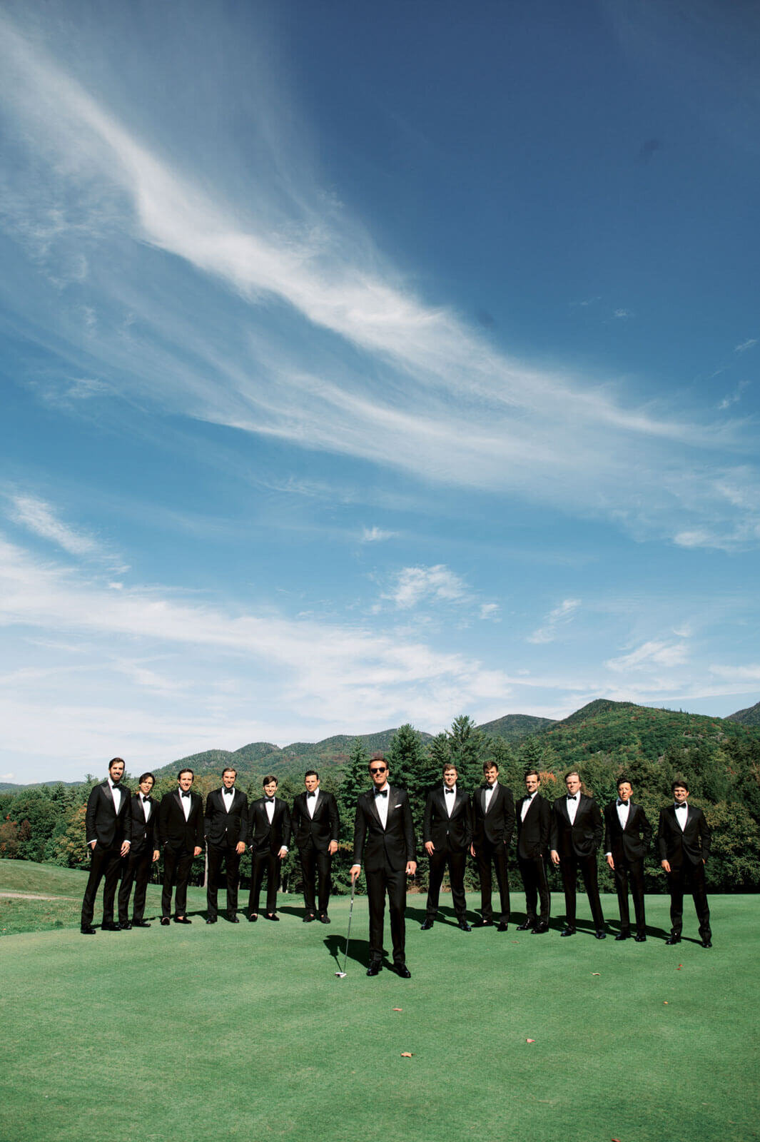The groom is with twelve other men in black suits at The Ausable Club, New York, with mountains and sky in the background.