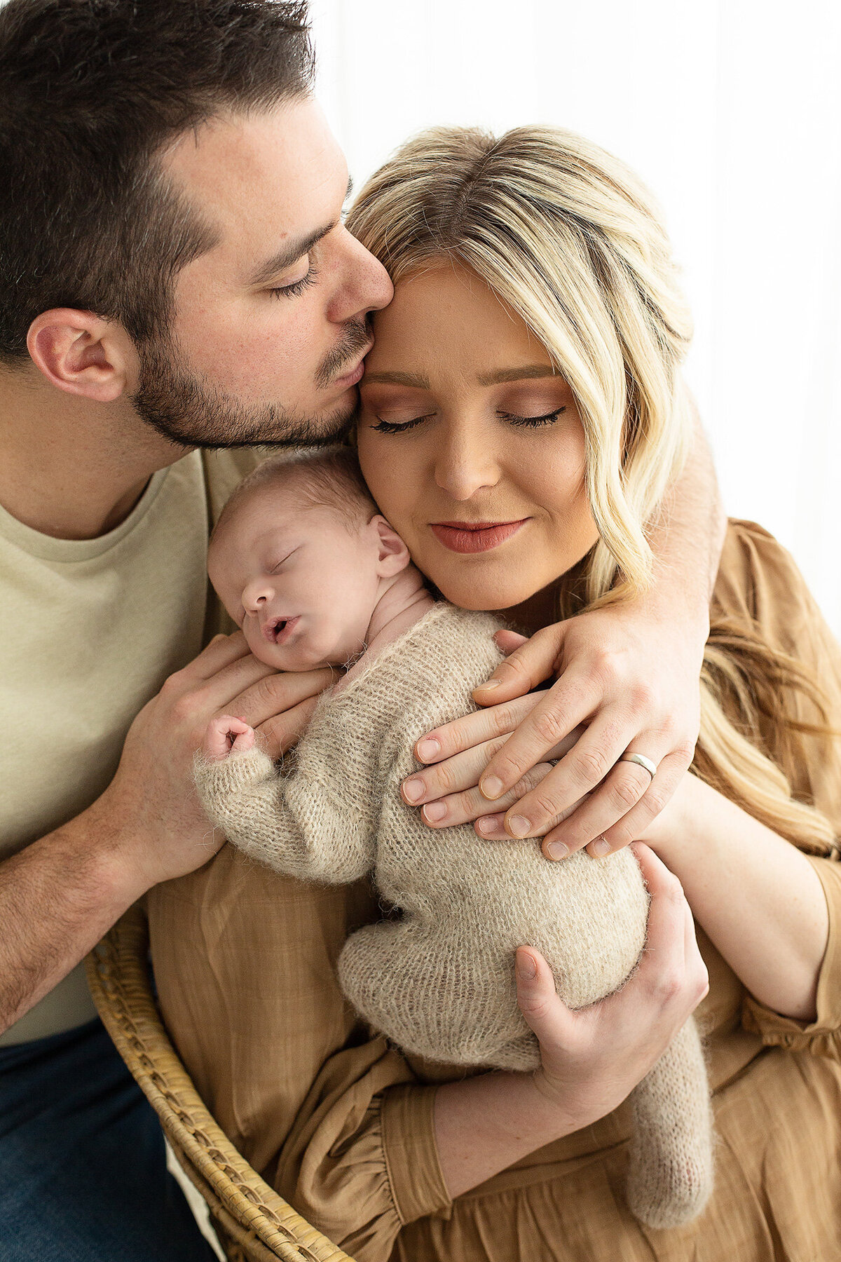 dayton-ohio-newborn-photographer-mom-sitting-in-chair-holding-baby-boy-on-shoulder-while-dad-kisses-her-on-forehead