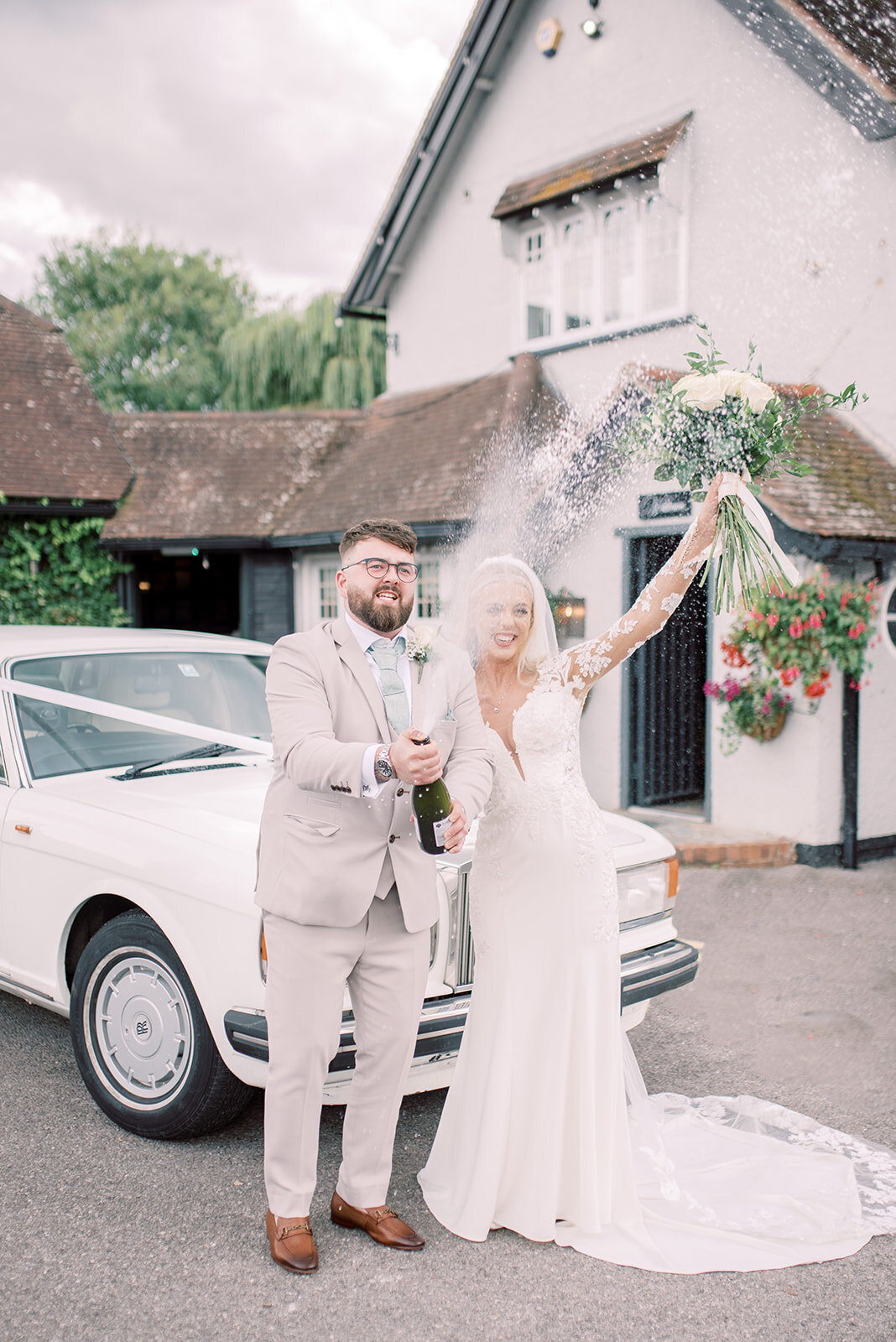 Bride and groom are standing in front of a rolls royce wedding car. spraying a bottle of champagne, the bride has her arm in the air and is smiling as they celebrate. The image is edited in a light and airy style.