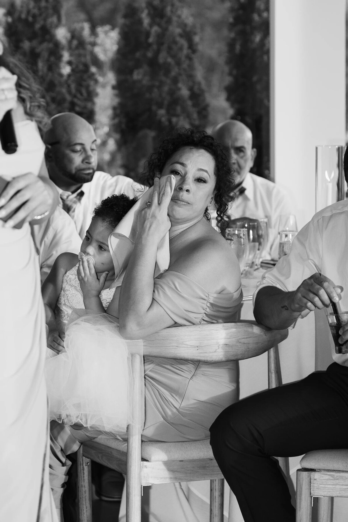 Rhode Island Wedding Photographers | Kelsey Sheehan Photography Timeless Rhode Island Weddings | A woman in a formal dress wipes her face while holding a child at a table during an emotional moment. Seated guests in the background. Black and white.