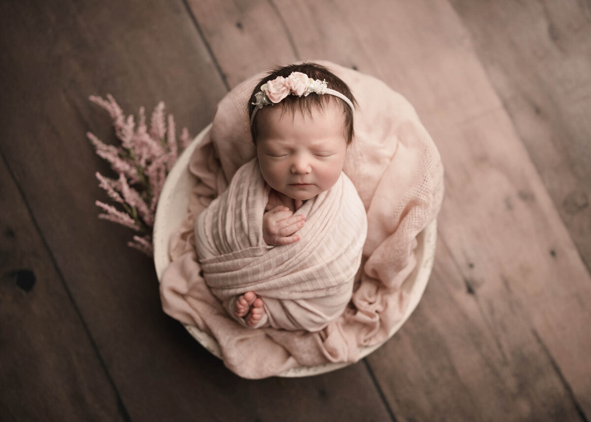 Aerial image of newborn photoshoot. Baby girl swaddled in blush fabric has her toes and fingers peeking out of the wrap. She is wearing a matching headband with small floral embellishments. Captured by best Lake Elsinore newborn photographer Bonny Lynn Photography.