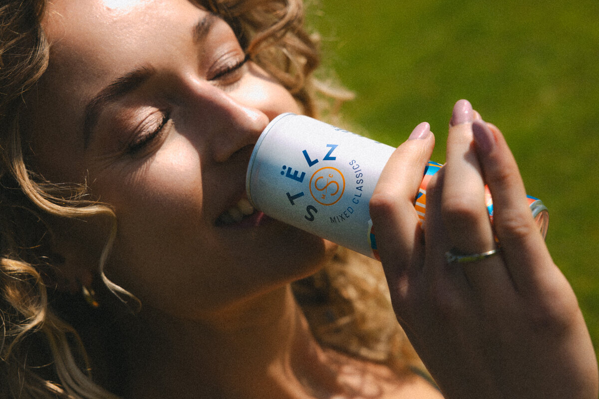 A close-up of a woman's face as she enjoys a sip of STËLZ Classic Spritz, her expression conveying satisfaction and delight. The soft focus and warm tones enhance the feeling of a perfect summer day. Photography by Bo Eppink.