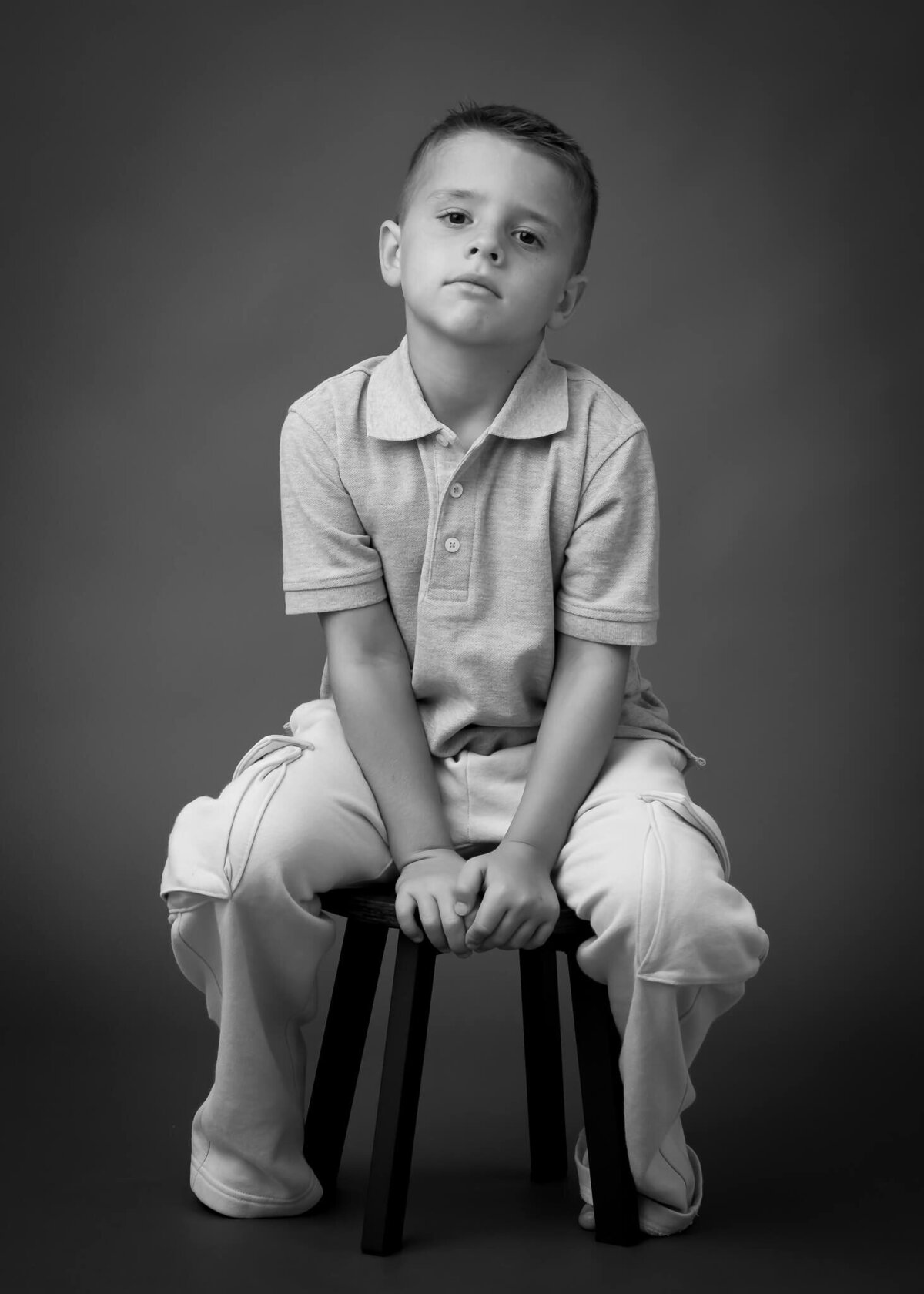 Moody black and white serious children's portrait of small boy with serious face sitting leaning forward on stool