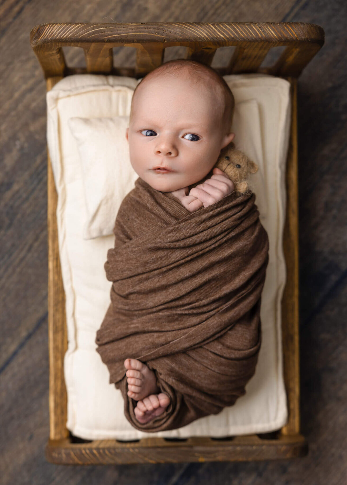 newborn baby with eyes open wrapped in brown fabric in a tiny wooden bed holding a tiny teddy bear