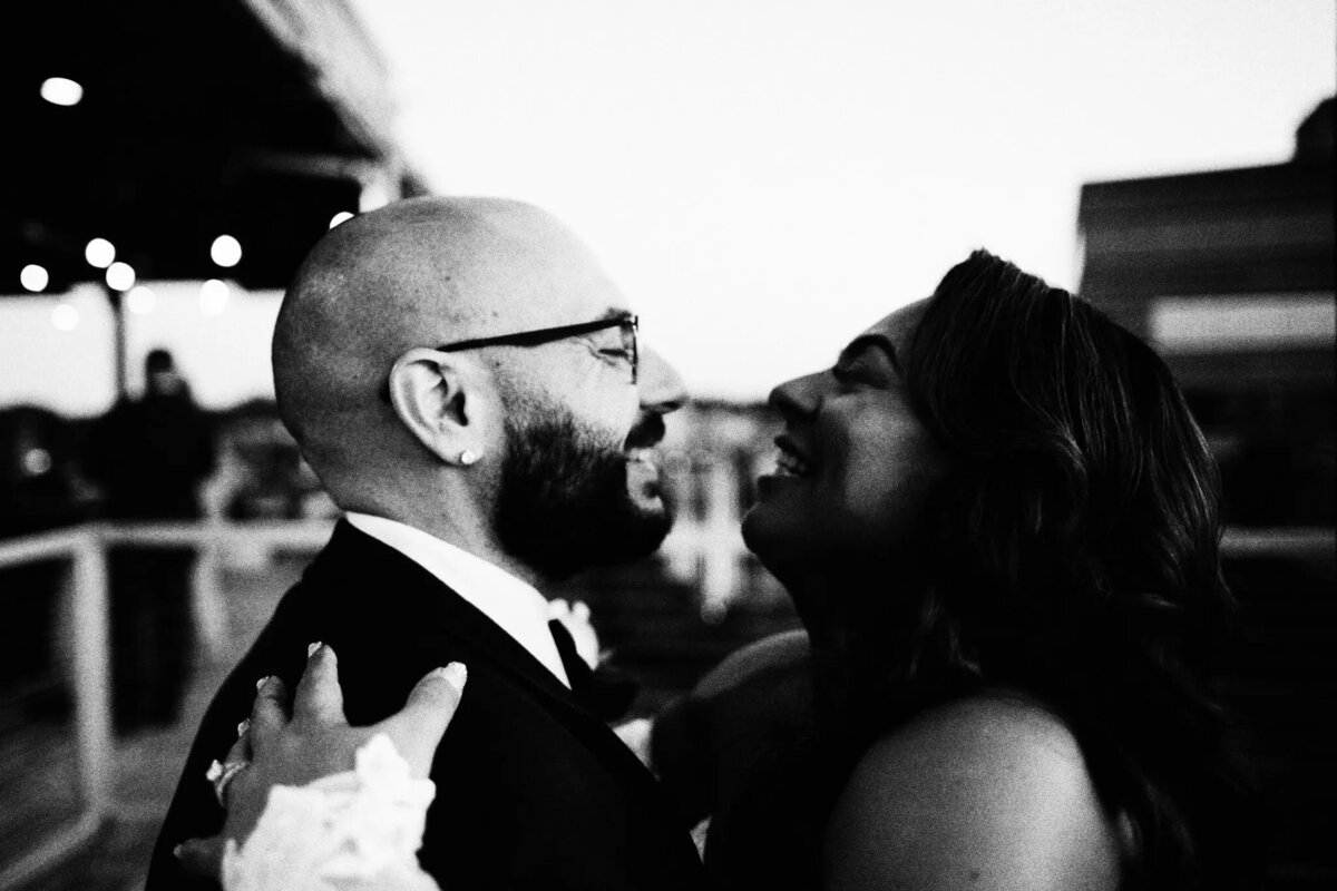 A joyful couple embraces and smiles at each other outdoors, captured in black and white by an NJ wedding photographer. The person on the left has glasses and a beard; the person on the right has long hair. The background is softly blurred.
