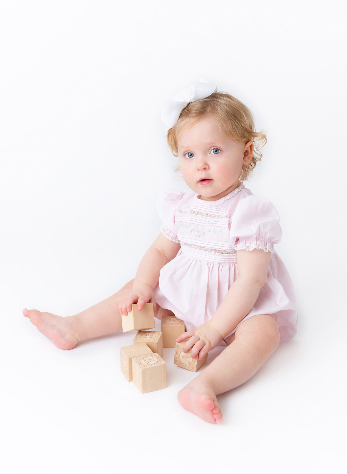Modern Heirloom baby portrait in Brooklyn — baby girl sitting softly with wooden alphabet blocks, wearing a classic pink dress and white bow, captured by NYC Heirloom photographer.