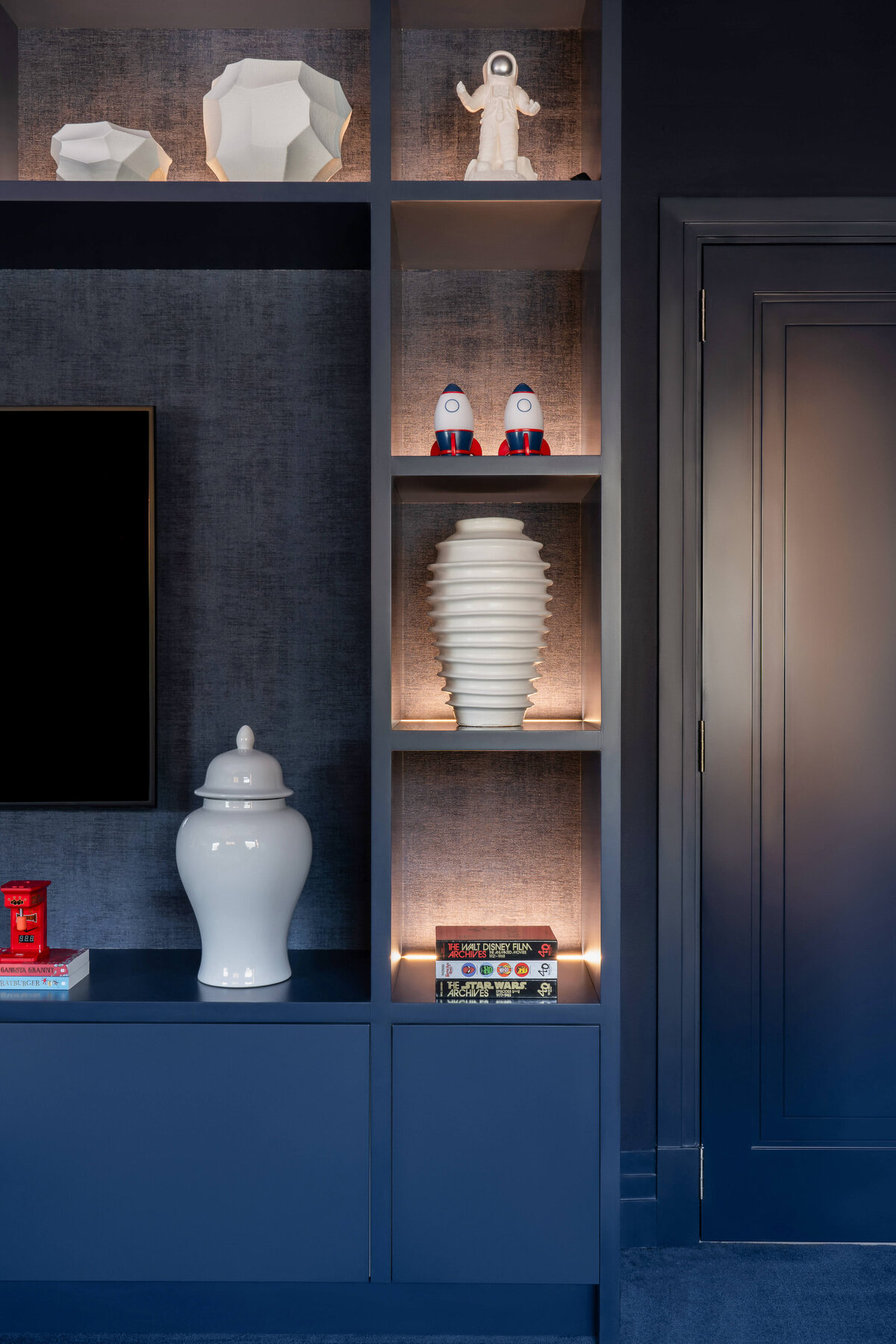 Built-in blue shelving around the television in a boy's outer space themed bedroom. The shelves are backlit to emit a soft glow and have white vases and children's toys.