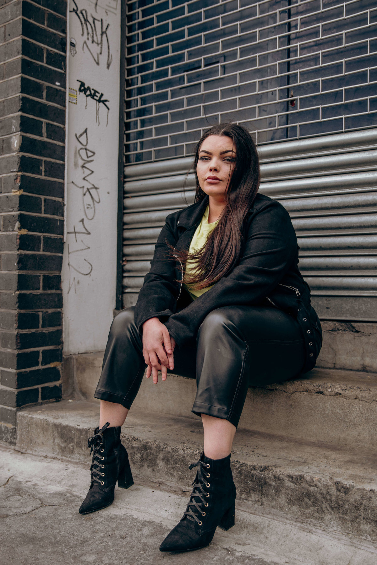 Tia wearing leather and a neon t shirt, sat on concrete steps infront of a metal shutter.