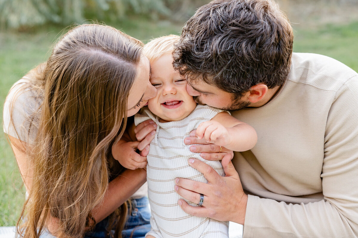 A mother and father gently kiss their giggling baby in a heartfelt moment captured in Eagle Mountain, Utah. This intimate family portrait showcases pure love and joy.