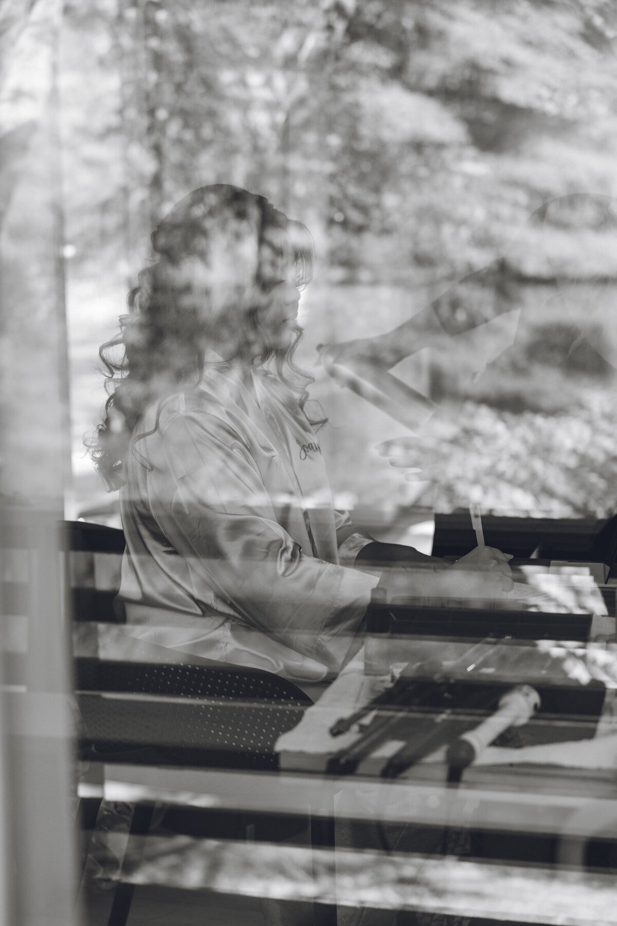 Bride getting ready, seen through a window reflection with trees in the background.
