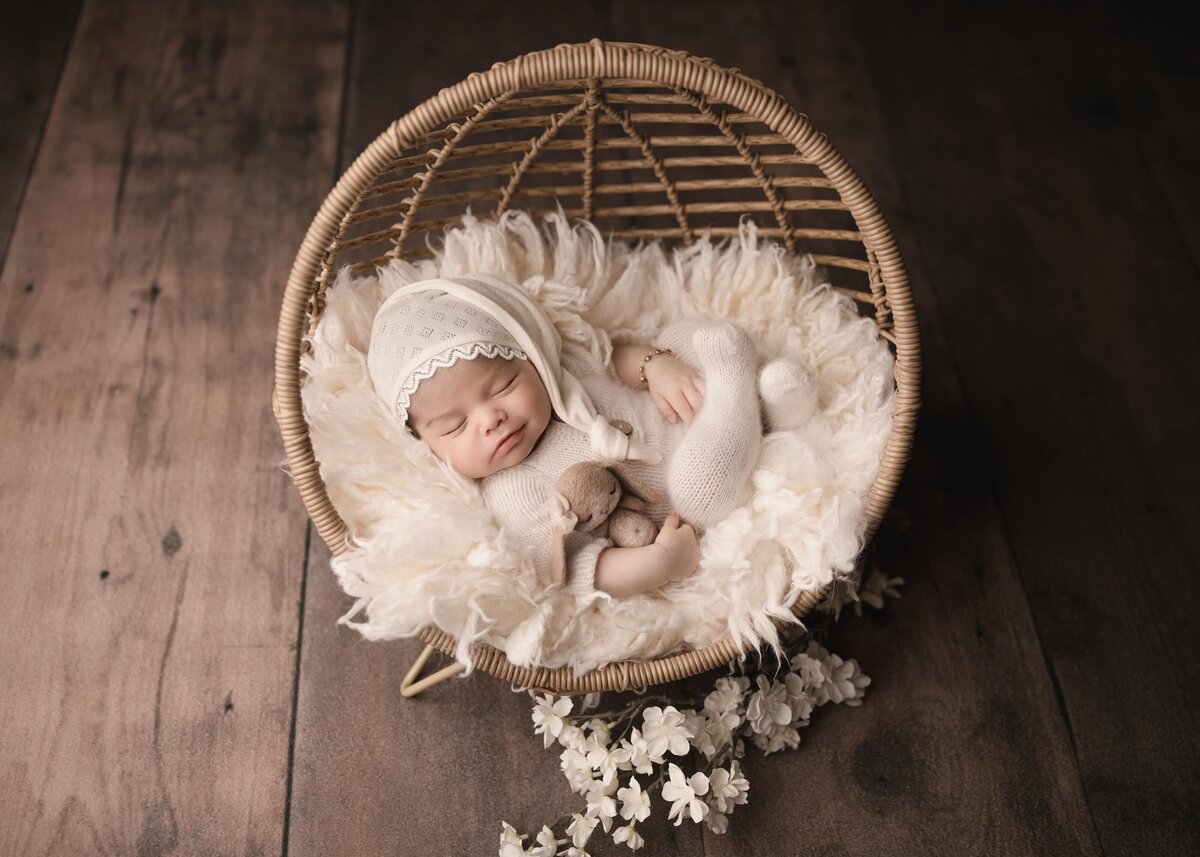 Menifee newborn baby girl photoshoot. Baby in a white knit onesie and long cap is laying in a miniature newborn is posed in a circle rattain chair. Her legs are folded on top of her. She has a little felt bunny under one arm. Captured by best Menifee newborn photographer Bonny Lynn Photography