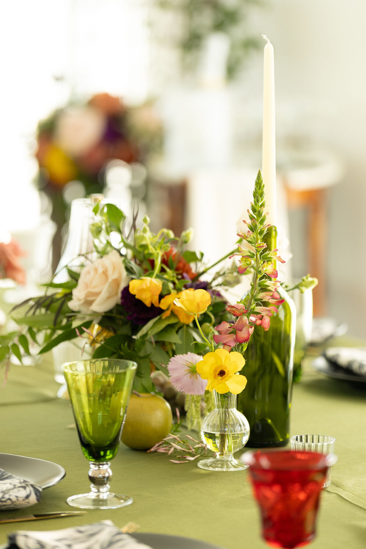 Small wedding bud vase arrangement with soft blush roses, yellow poppies, and wild garden blooms styled on an olive-green reception table with colored glassware.