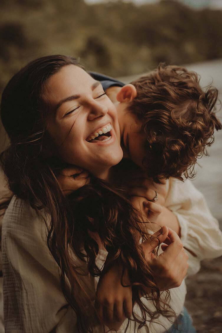 A son stands behind his mother while giving her big hugs and she laughs