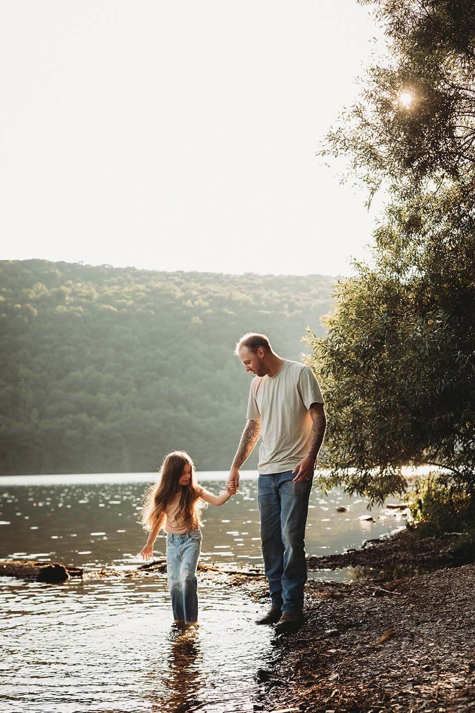 A dad holds his daughters hand while she walks barefoot in the water at the lake. Her jeans are wet and they are laughing.