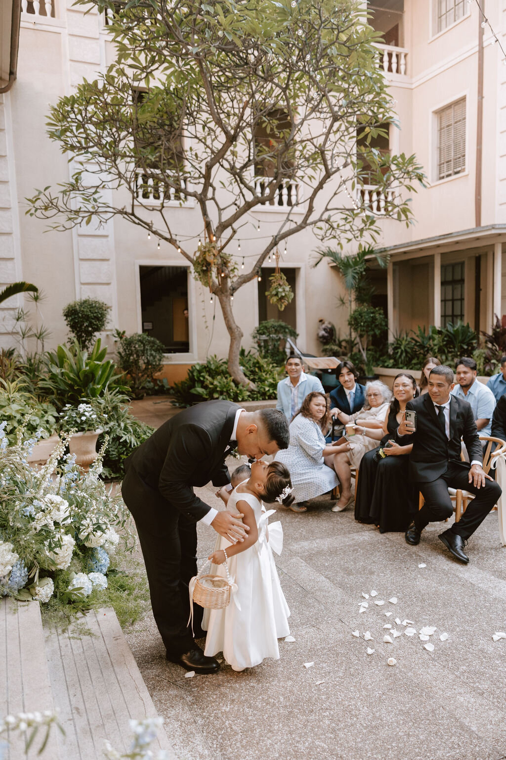 Flower girl looking up to kiss groom while guests are watching and tree in the background