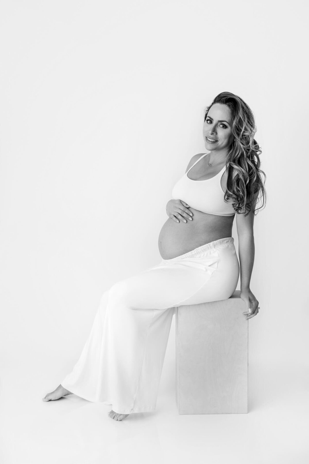 A pregnant woman sits on a rectangular block against a white background, exuding elegance and serenity. She is wearing a white crop top and flowing white pants that accentuate her baby bump. Her long, wavy hair cascades over her shoulder as she smiles softly at the camera. One hand rests on her belly, while the other supports her position on the block.