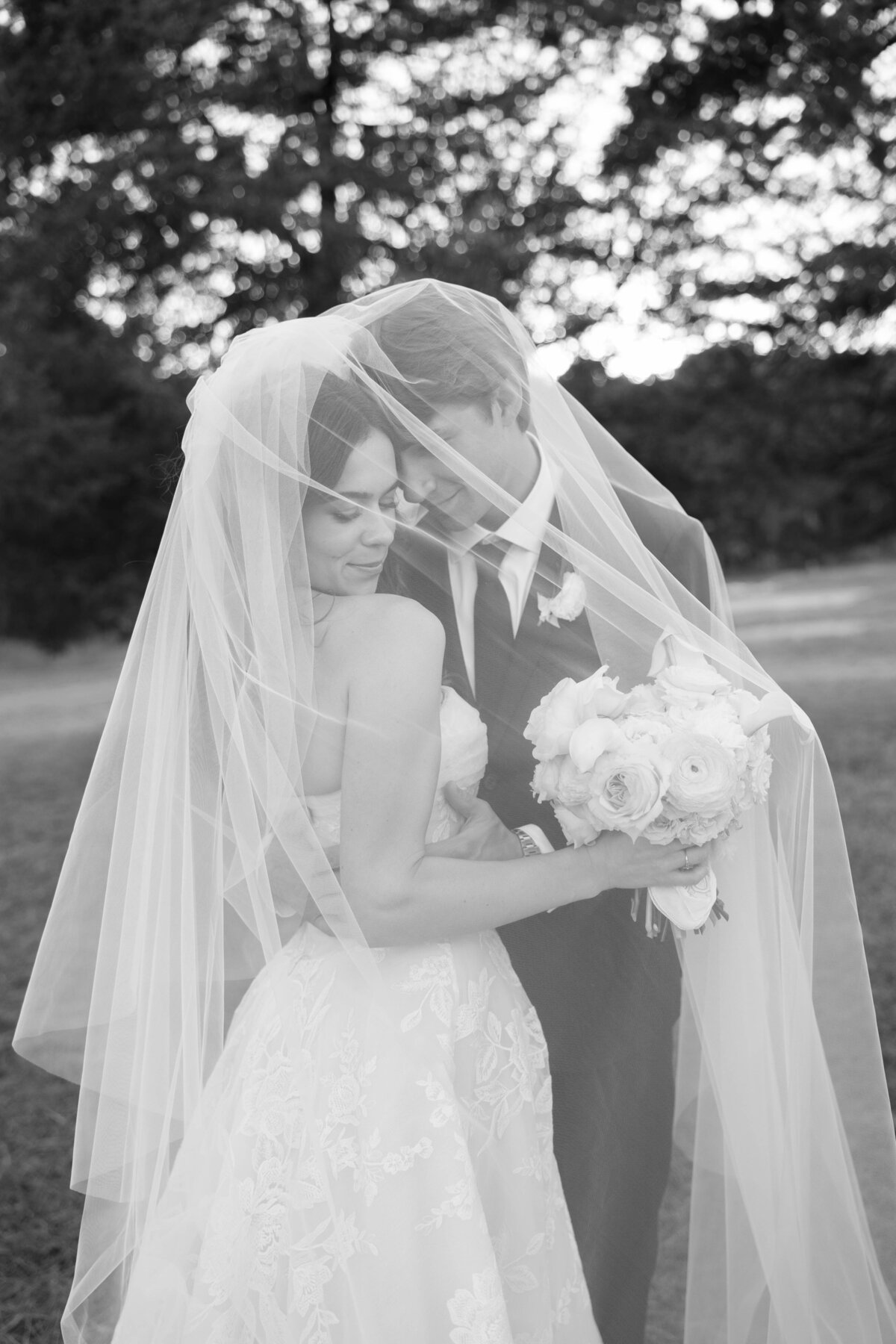 Romantic black-and-white photo of bride and groom embracing under the wedding veil with a soft, airy bouquet of white and blush florals arranged by an Arkansas wedding florist.