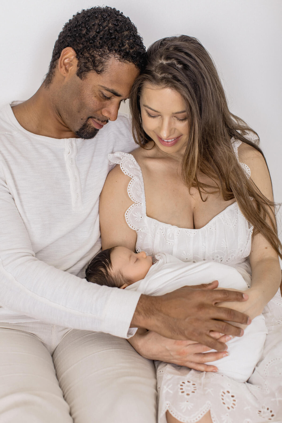 A couple lovingly cradles a sleeping newborn wrapped in a white blanket. They are sitting close together, looking at the baby with affection. The soothing colors and soft lighting create a warm and tender atmosphere.
