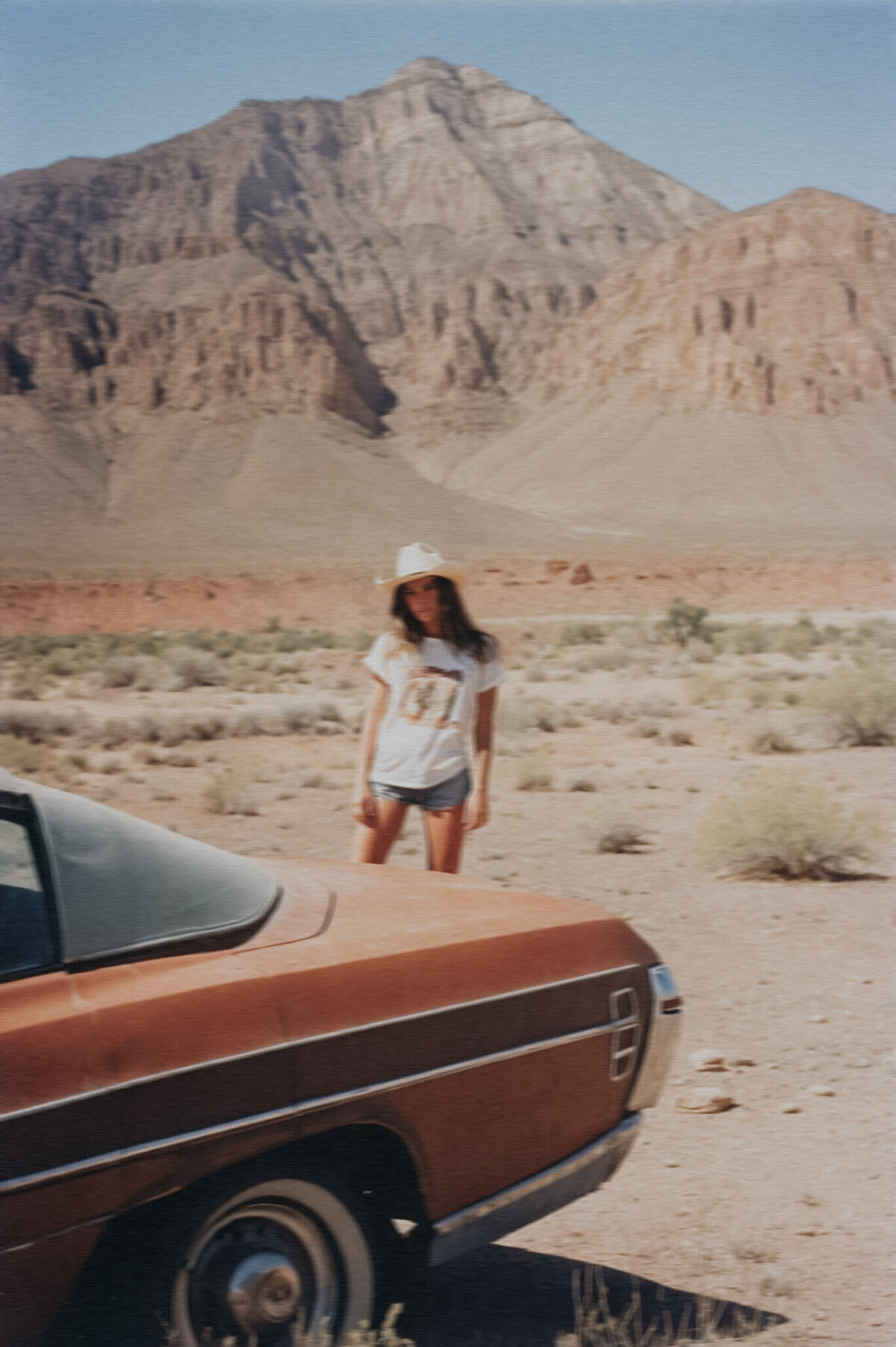 A person in a white t-shirt and hat stands beside a vintage car in a desert landscape with a large mountain in the background, capturing the timeless beauty perfect for Sweet Pea Ink Creative's Showit web design inspiration.