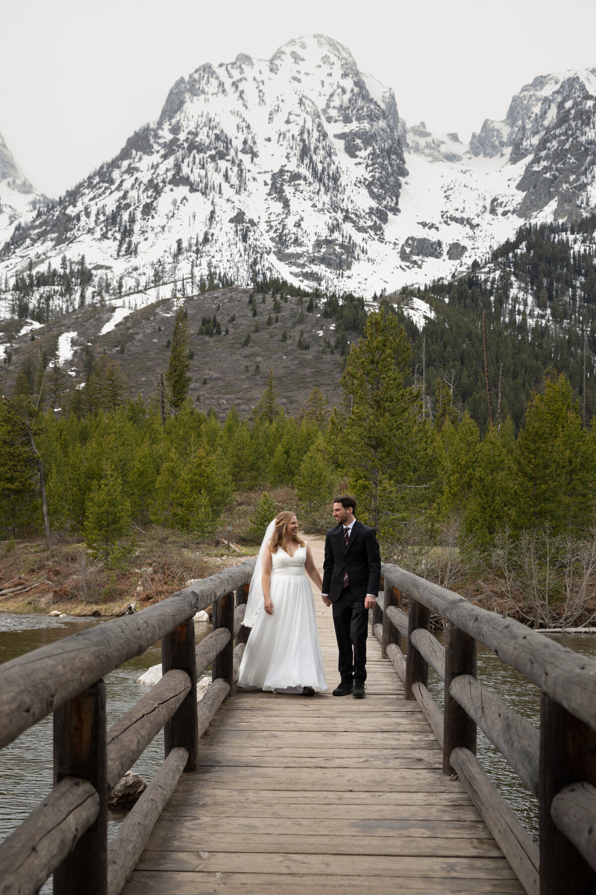 A bride and groom walk hand in hand along a wooden bridge while their Wyoming elopement photographer takes their photo.