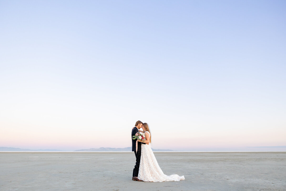 Bride and groom stand close together, holding a bouquet as they kiss under a soft sunrise sky.