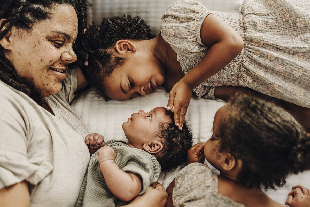 Newborn baby looking up at his big sister, surrounded by Mom smiling at him and other sister looking on: Statesboro lifestyle newborn photography