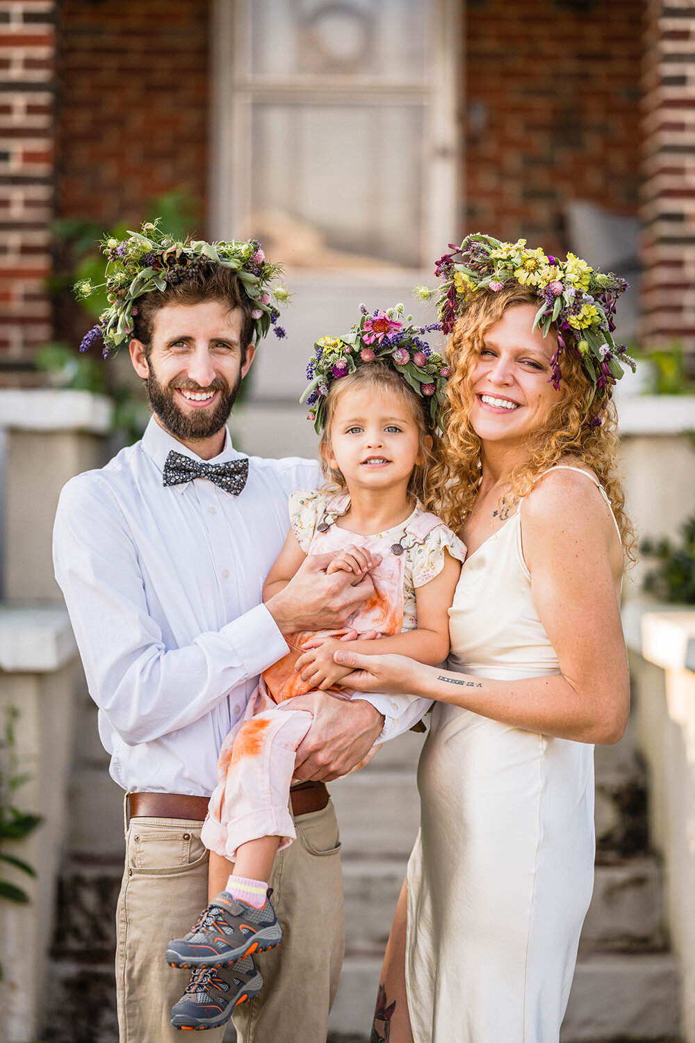 A family of three stand outside of their home for a portrait before heading to their ceremony spot on their elopement day. The trio are wearing handmade flower crowns and are wearing casual wedding attire.