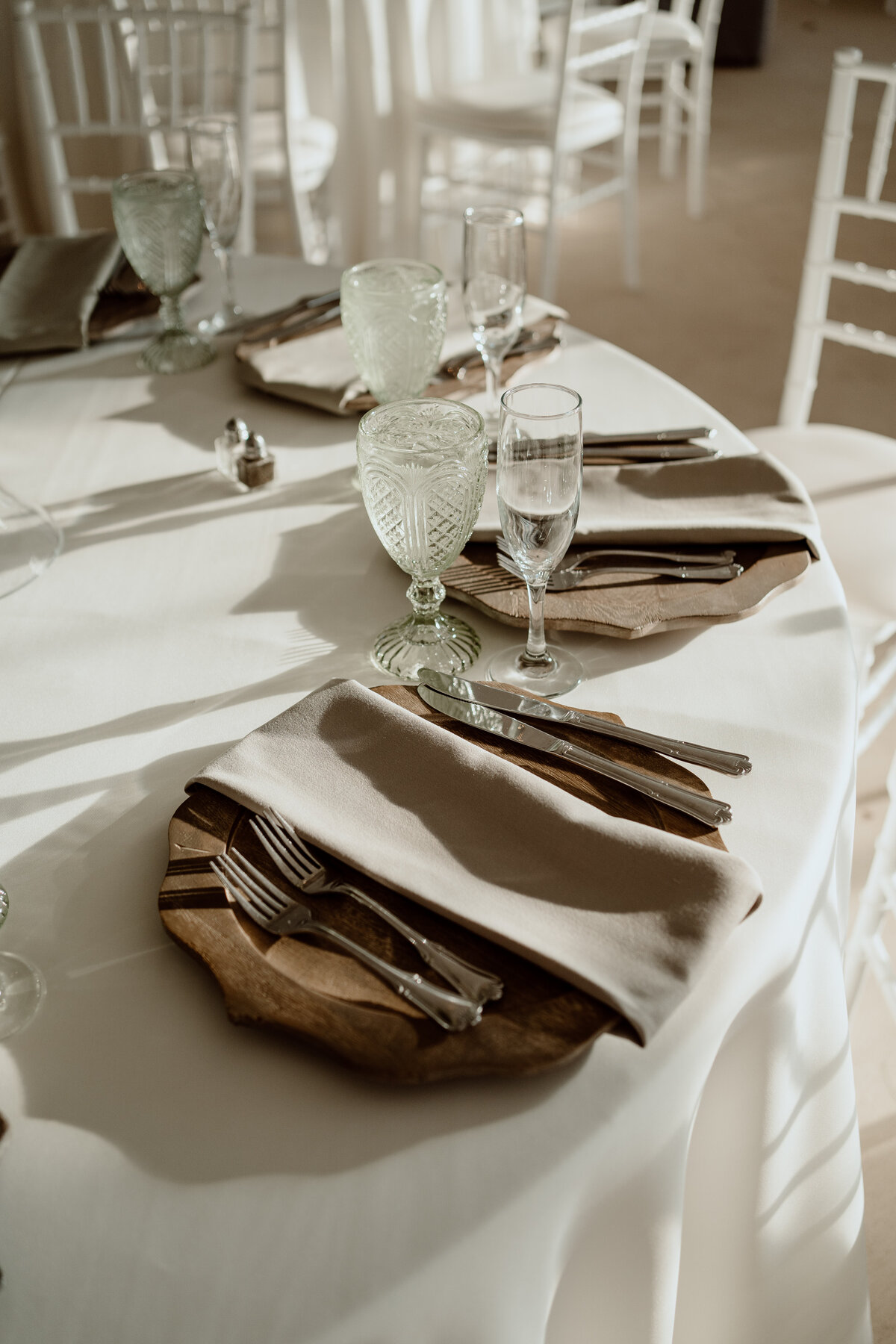 Close-up of an elegant wedding reception table setting, featuring wooden chargers, beige napkins, vintage glassware, and silver cutlery on a white tablecloth. The natural light creates a warm and inviting atmosphere.