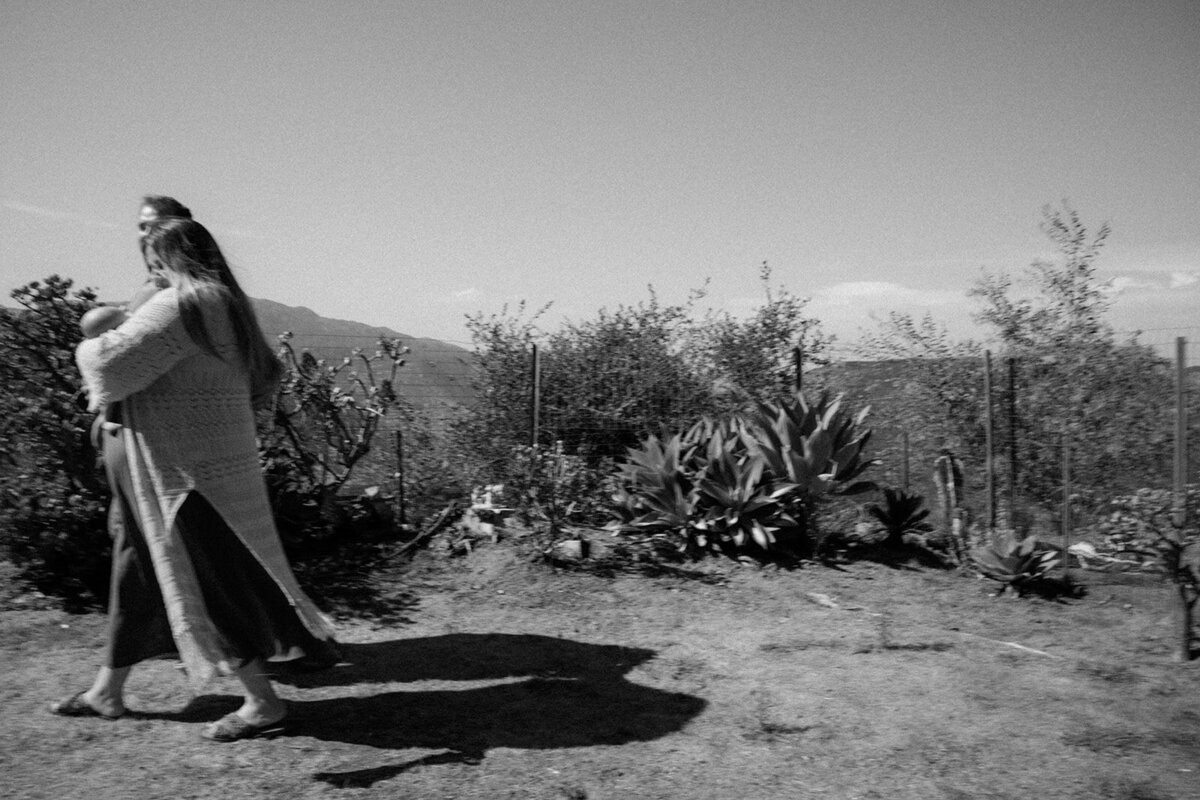 Mother and father hold infant while walking past cactus in the San Diego terrain. 