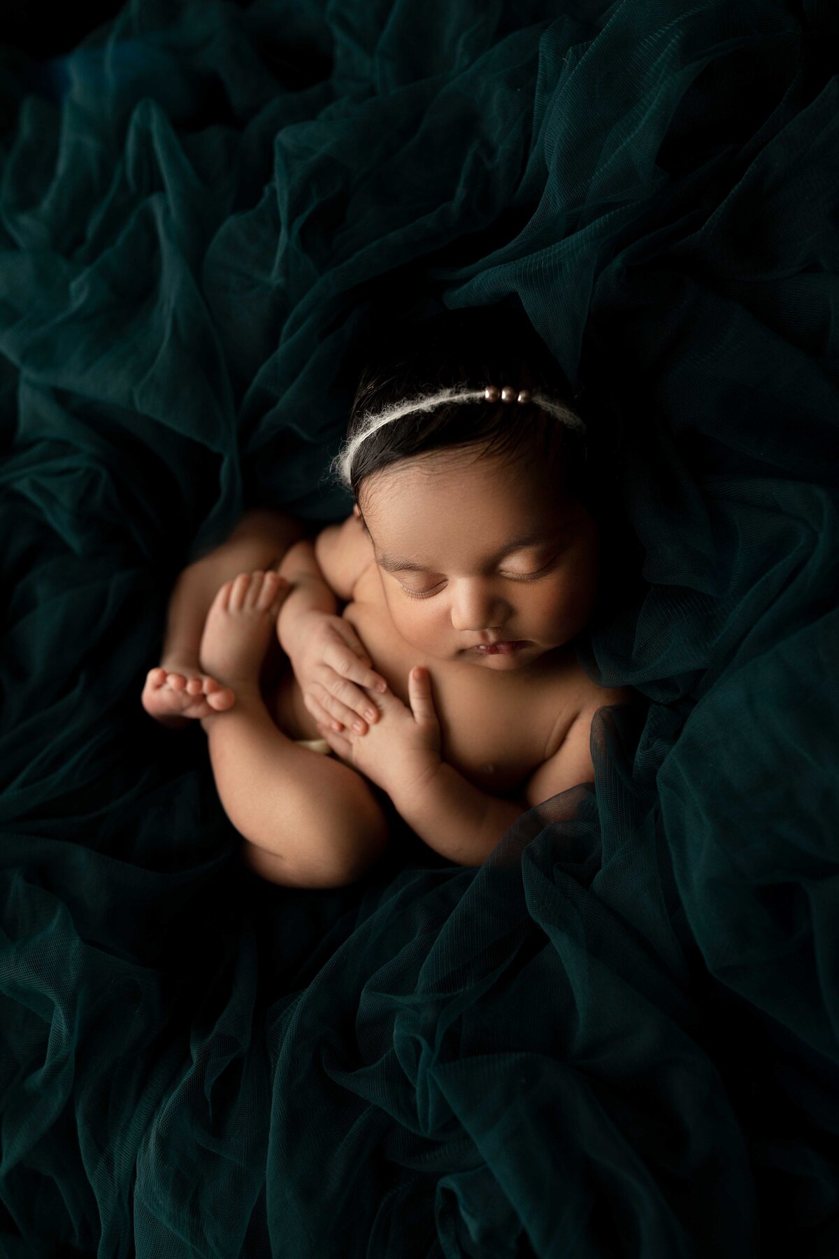 Top Newborn  Studio Photographer in London, Ontario- Baby curled up sleeping on emerald green velvet with delicate headband. Hands crossed on chest.