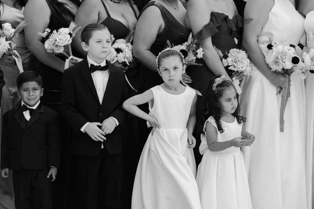 A black and white detail shot of four child attendants at a wedding in Dallas, Texas. The two boys are both wearing tuxedos while the two girls are wearing light dresses.  One boy looks slightly distressed while the other looks pensive. One of the girls is looking questioningly at the camera while the other watches intently off to the side. The four children are backed by the bride and the bridesmaids.