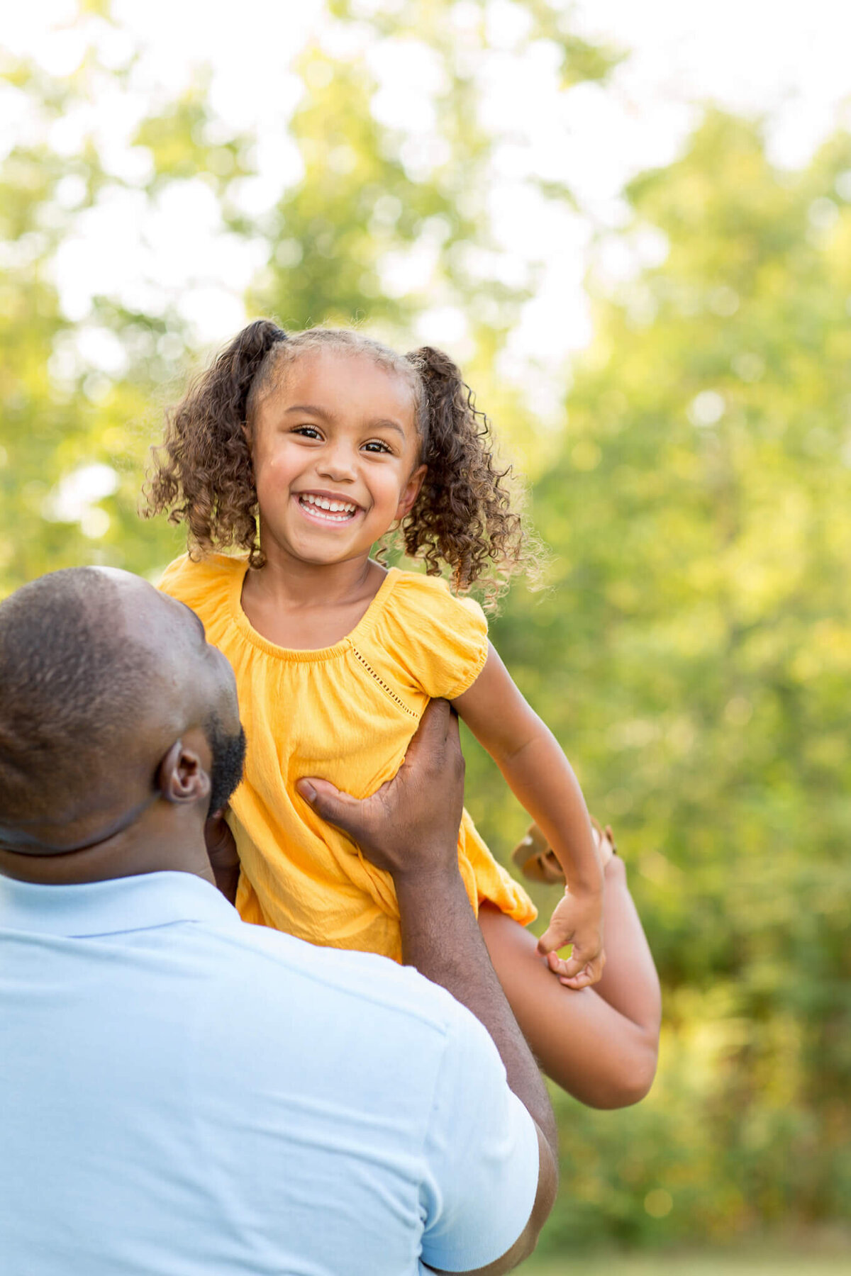 A man in a blue shirt joyfully lifts his smiling daughter, her yellow dress swaying as her pigtails bounce. Captured by a Fayetteville NC family photographer, they are surrounded by lush green trees, basking in the sunshine of a perfect day outdoors.