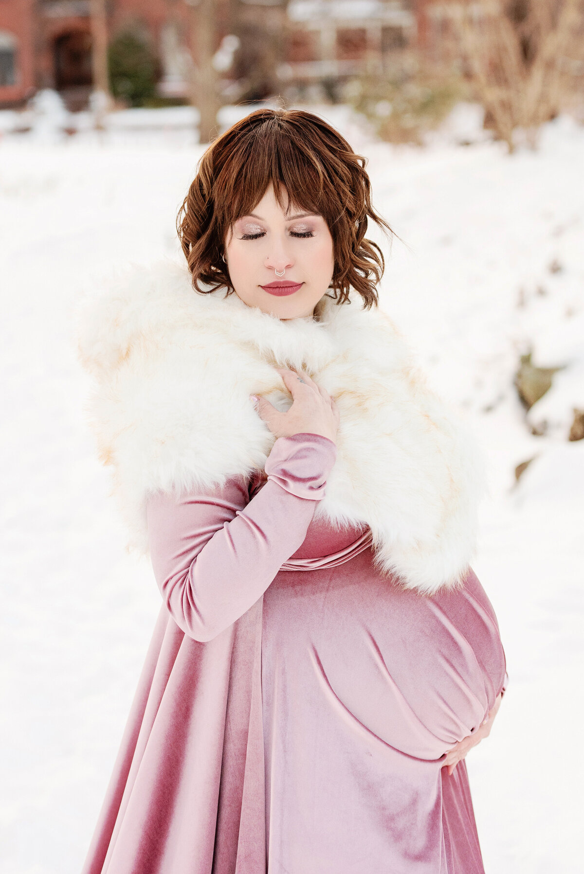 st-louis-maternity-photographer-expecting-mother-posed-holding-belly-in-pink-velvet-gown-wth-white-fur-against-white-snowy-background