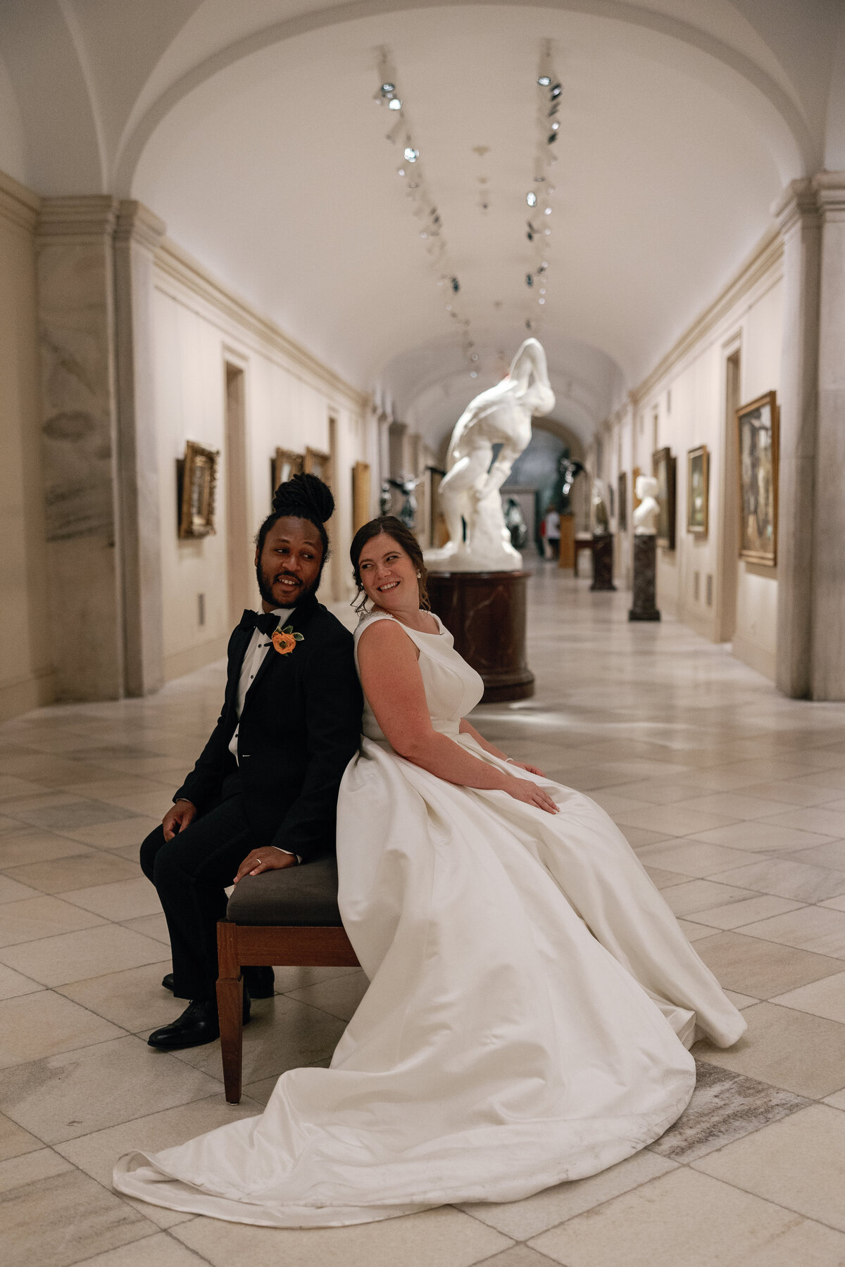 A bride and groom sit on a bench inside the National Portrait Gallery in front of a marble sculpture. 