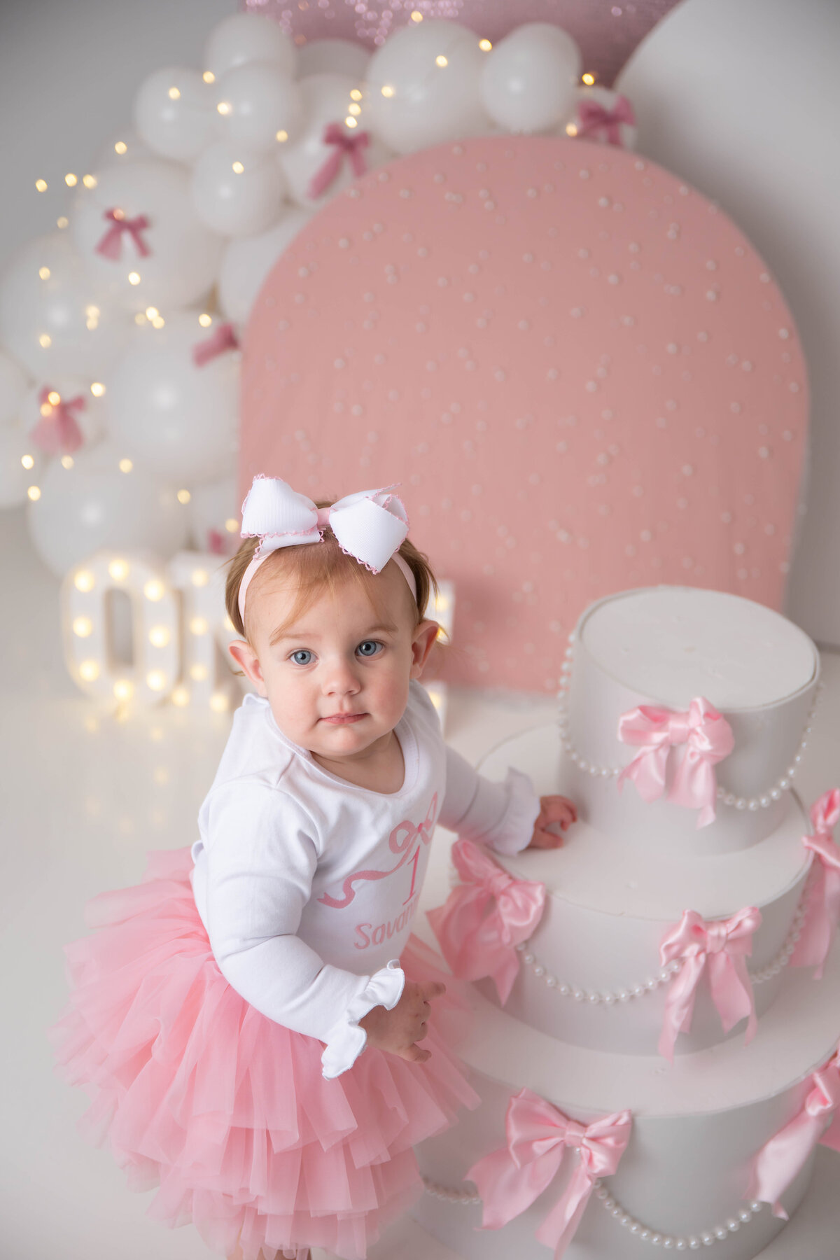Baby glances up as she has one hand on a cake prop with half of a led sign showing behind her. 
