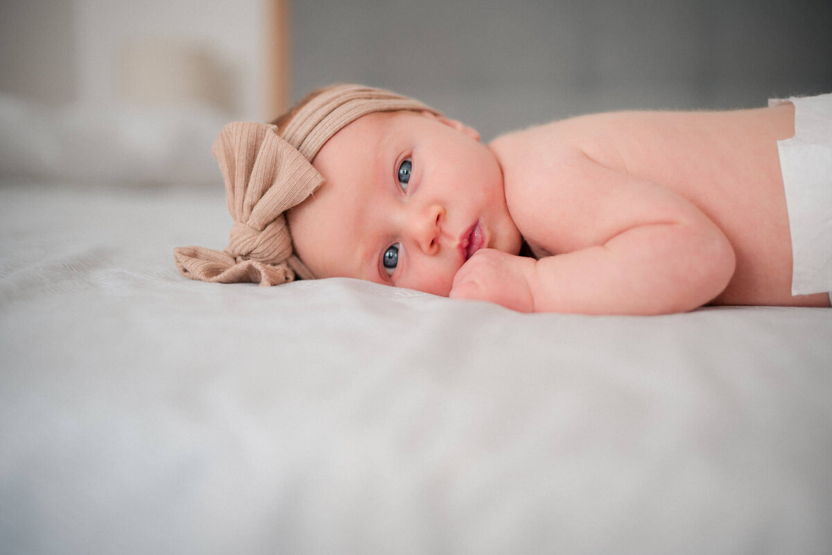 Newborn baby wearing a headband with a bow, lying awake on a bed and gazing toward the camera.