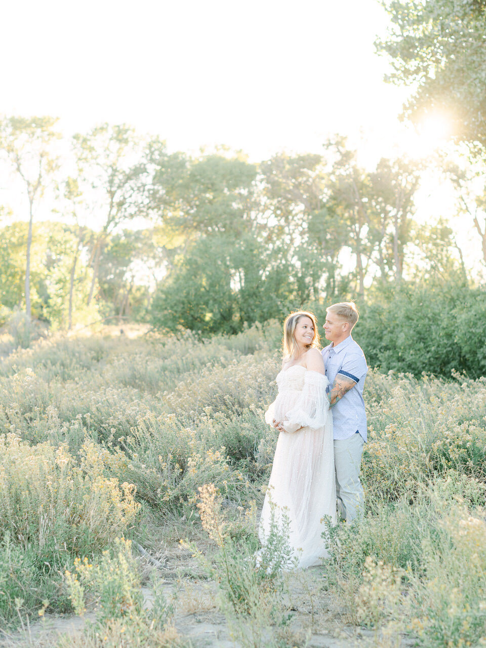 A couple embracing in a sunlit meadow during a maternity session, with the mother-to-be in a flowing off-the-shoulder gown, captured in an artful editorial style by Elizabeth Kane Photography—serving Philadelphia, New York, and DC.