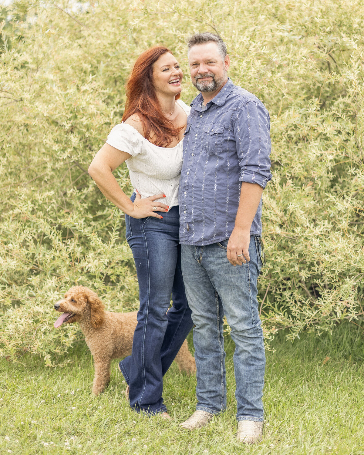 Mom and Dad are standing in front of a bush with their light brown dog at their feet.  Mom is looking at Dad laughing and Dad is looking at the camera smiling.  Dad is wearing blue jeans, blue and white stripped button up shirt, and has short pepper hair and beard.  Mom is wearing blue jeans, short sleeve white shirt, and has long red hair. 