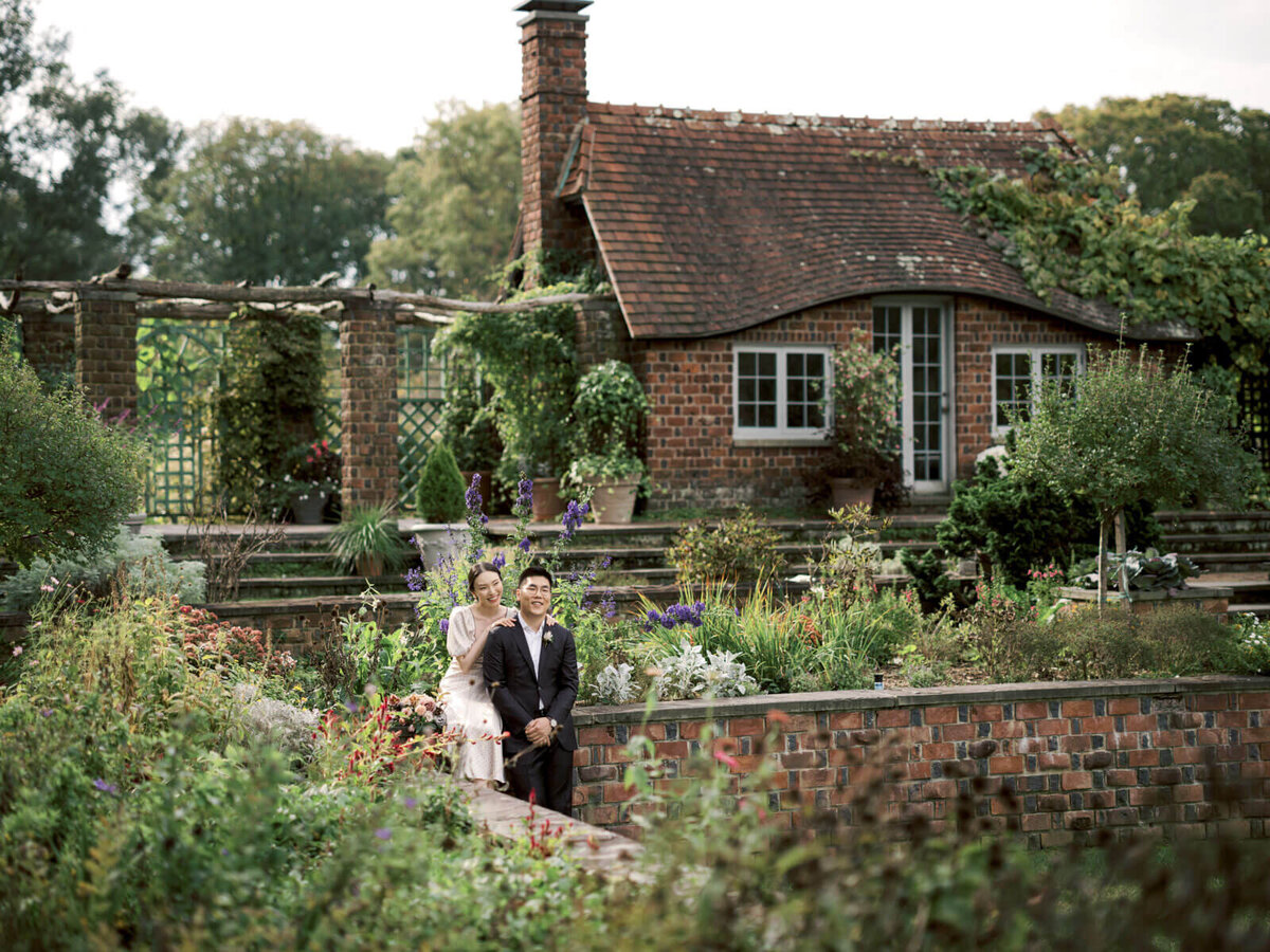 The engaged couple is in front of a red brick cottage, amidst a lovely garden, at Planting Fields Arboretum, NY. Image by Jenny Fu Studio
