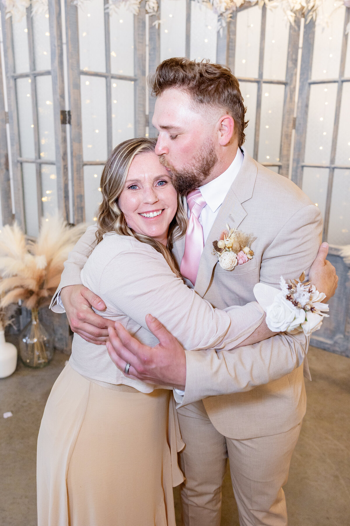 The groom kisses his mother on the head as they embrace, capturing a touching mother-son moment.